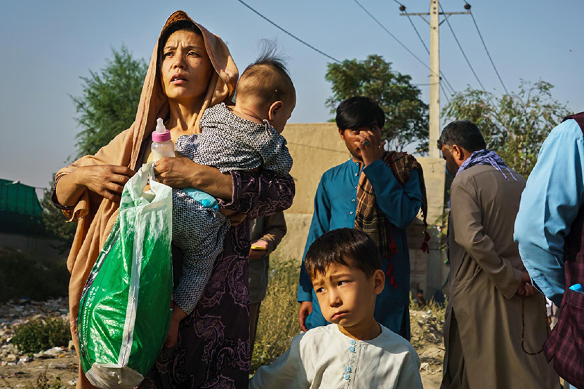 A woman holds a child and a bag while another child and men stand nearby outdoors.