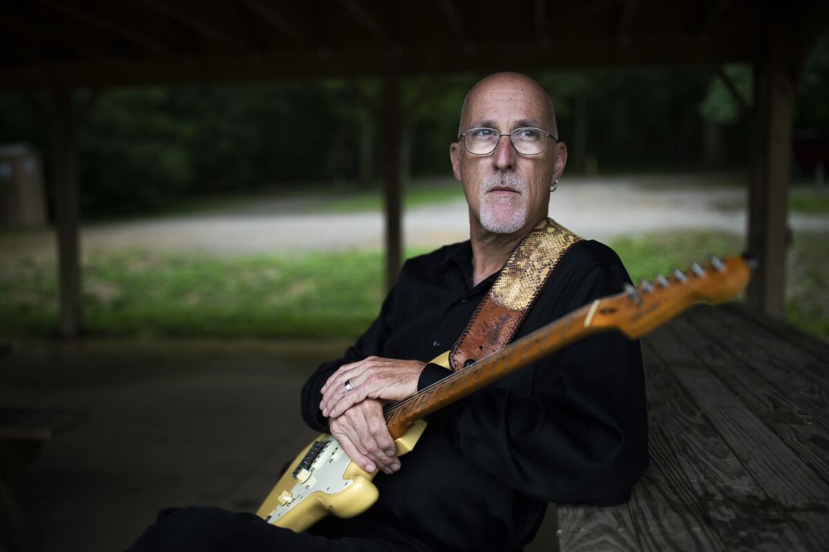 A man with an electric guitar sits and leans against a picnic table