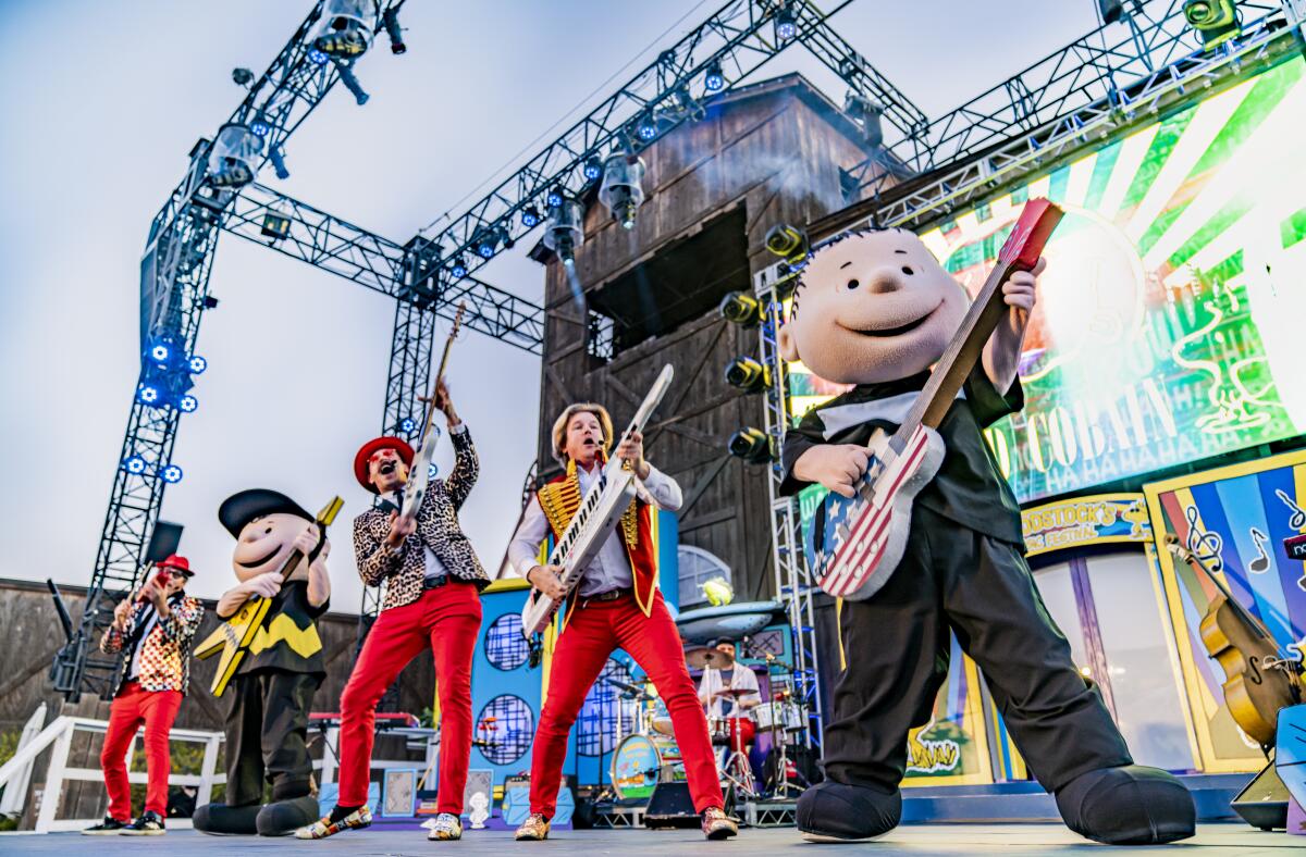 Men in red pants and colorful jackets play musical instruments onstage alongside people costumed as Peanuts characters.