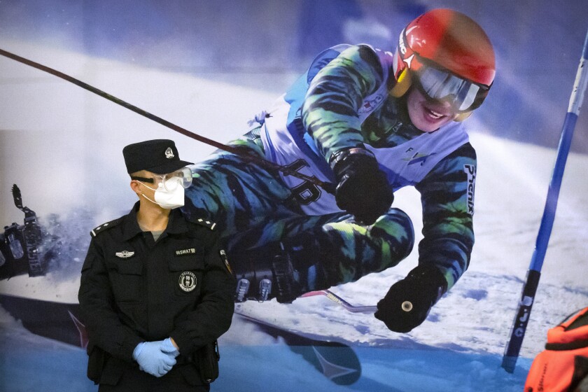ARCHIVO - Un agente de policía con mascarilla y gafas protectoras contra el COVID-19, de pie ante un cartel con la imagen de una esquiadora en una estación de tren en Zhangjiakou, en la provincia norteña china de Hebei, el viernes 26 de noviembre de 2021. (AP Foto/Mark Schiefelbein, Archivo)