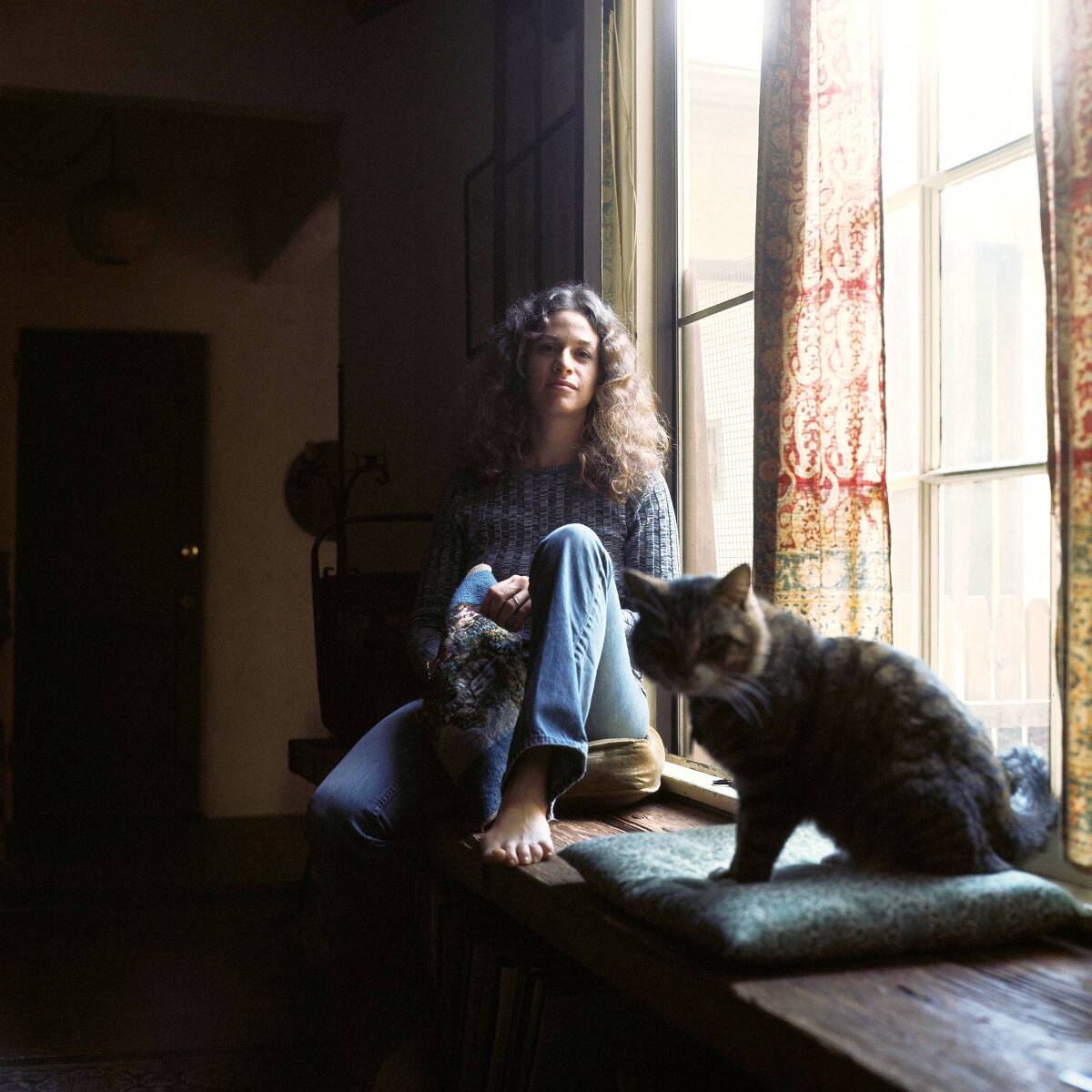 Carole King sits next to a window, with a cat.