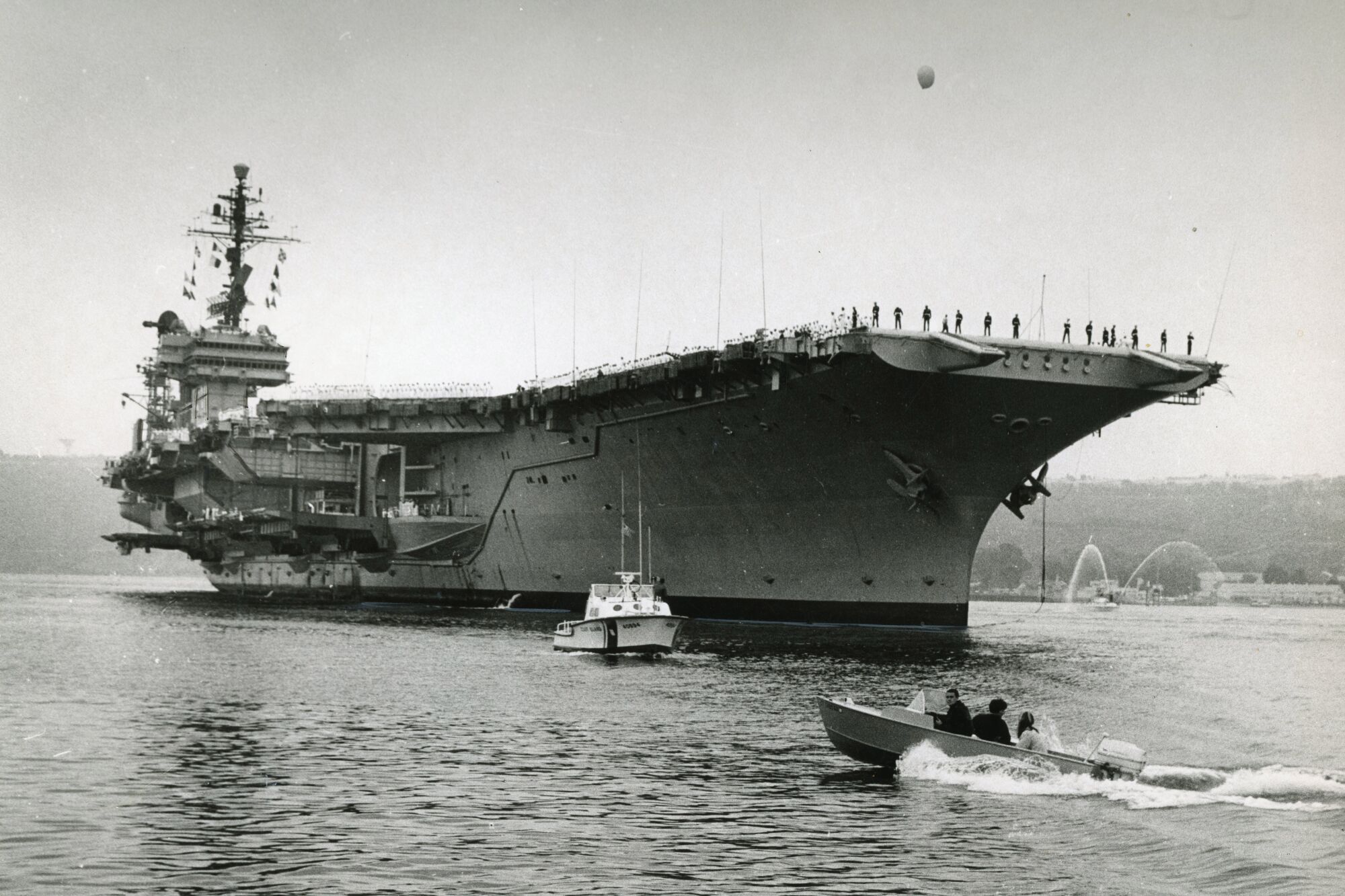 The aircraft carrier Kitty Hawk eases past Point Loma as an outboard motorboat roars beneath her bow in 1968.