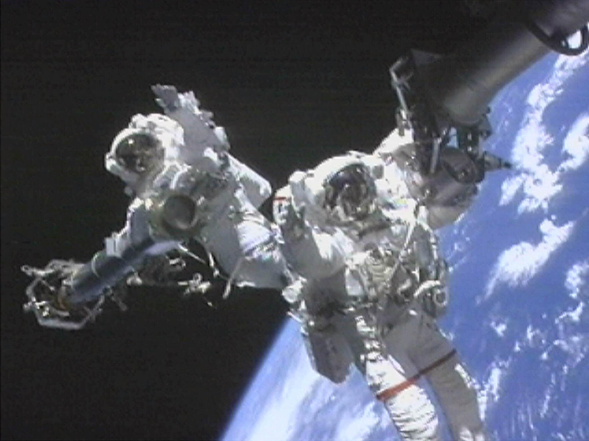 Two astronauts in spacesuits on a space walk, the Earth visible behind them