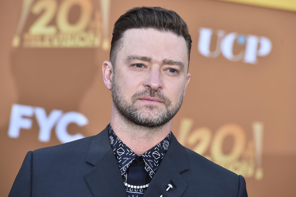 Justin Timberlake wearing a printed shirt, black tie and blazer in front of a dull orange backdrop