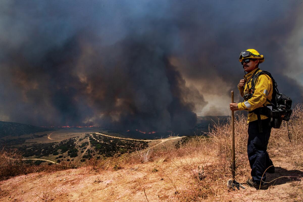 A firefighter stands on a hillside as a fire burns and smoke billows in the distance.