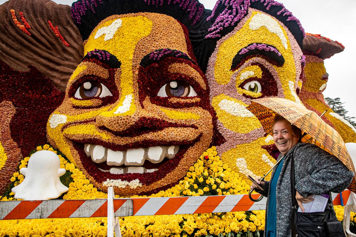 a woman with an umbrella smiles in front of an intricate yellow face sculpture made of real flowers