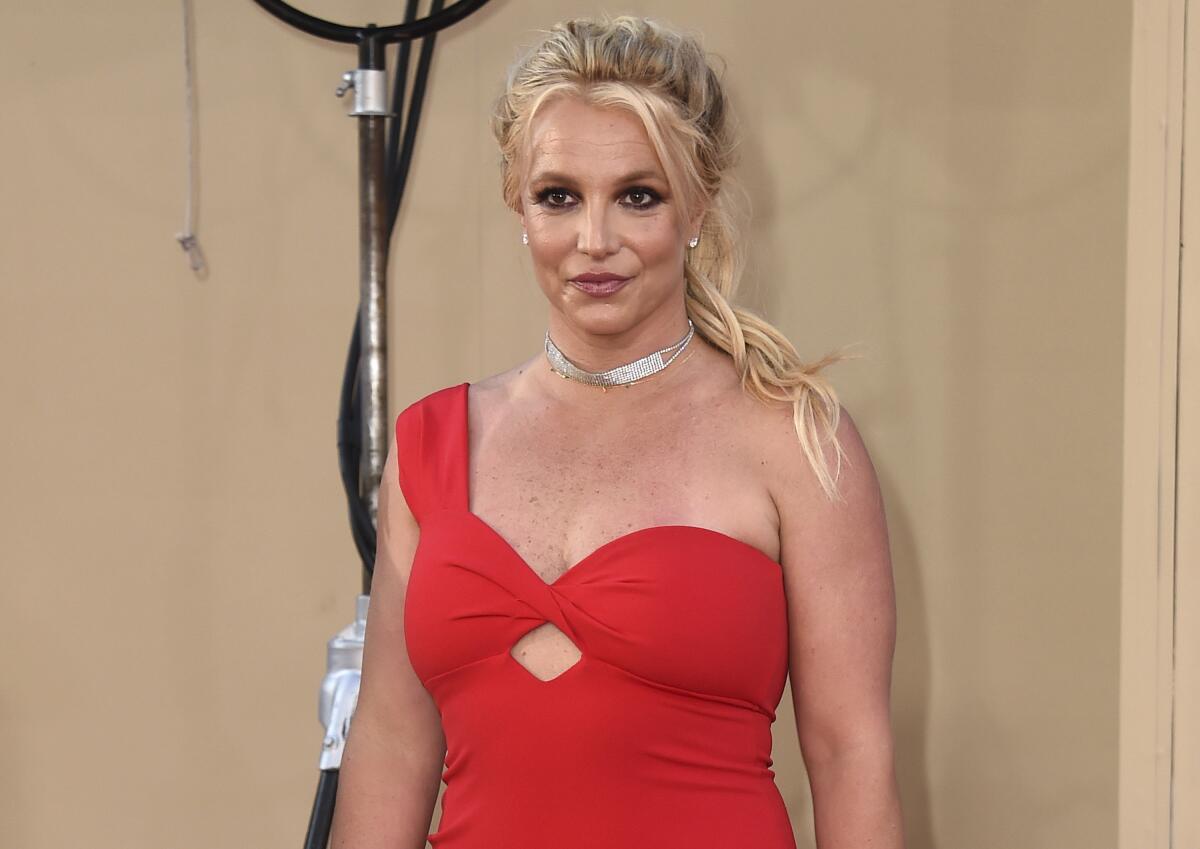 Britney Spears, wearing a one‑shoulder red dress, poses for photos.