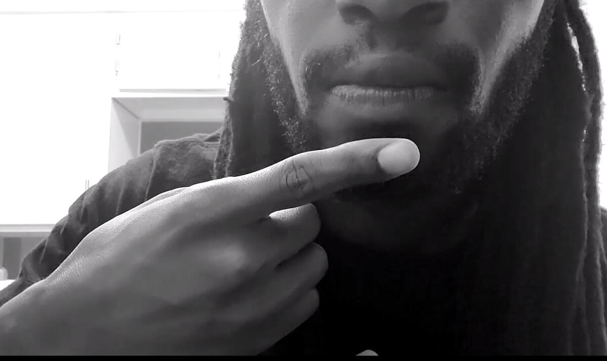 A close-up of actor Malcom Stokes taps his chin, making the ASL symbol for "say it" or "speak it."