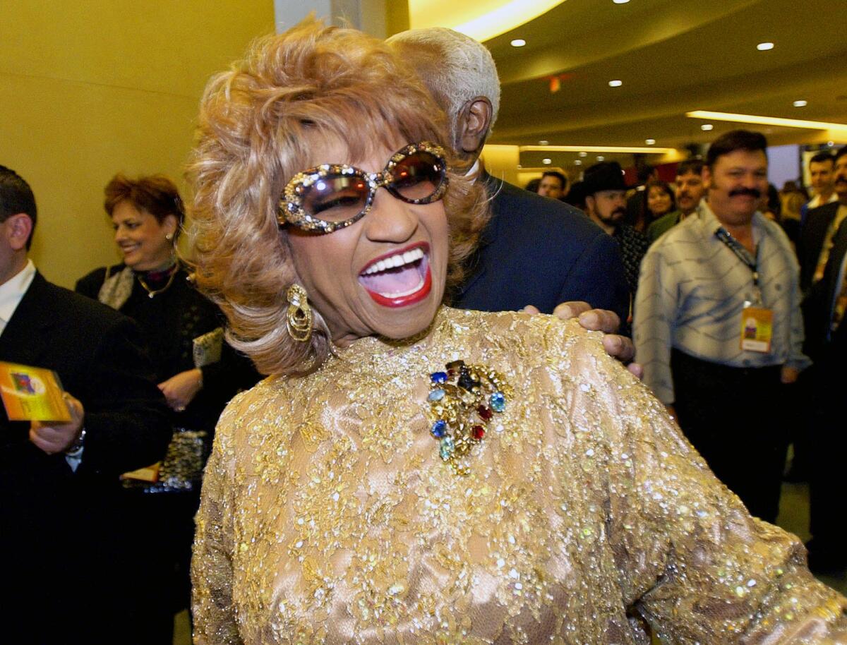 A woman smiles with mouth open, with blond hair, large sunglasses and a gold, shimmering dress
