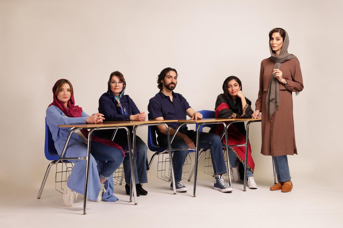 The cast of "English" at the Old Globe sit together at four school desks with a teacher standing to the right.