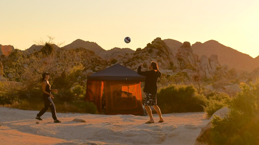 Two people play with a ball outside a tent backed by desert plants and mountain crags.