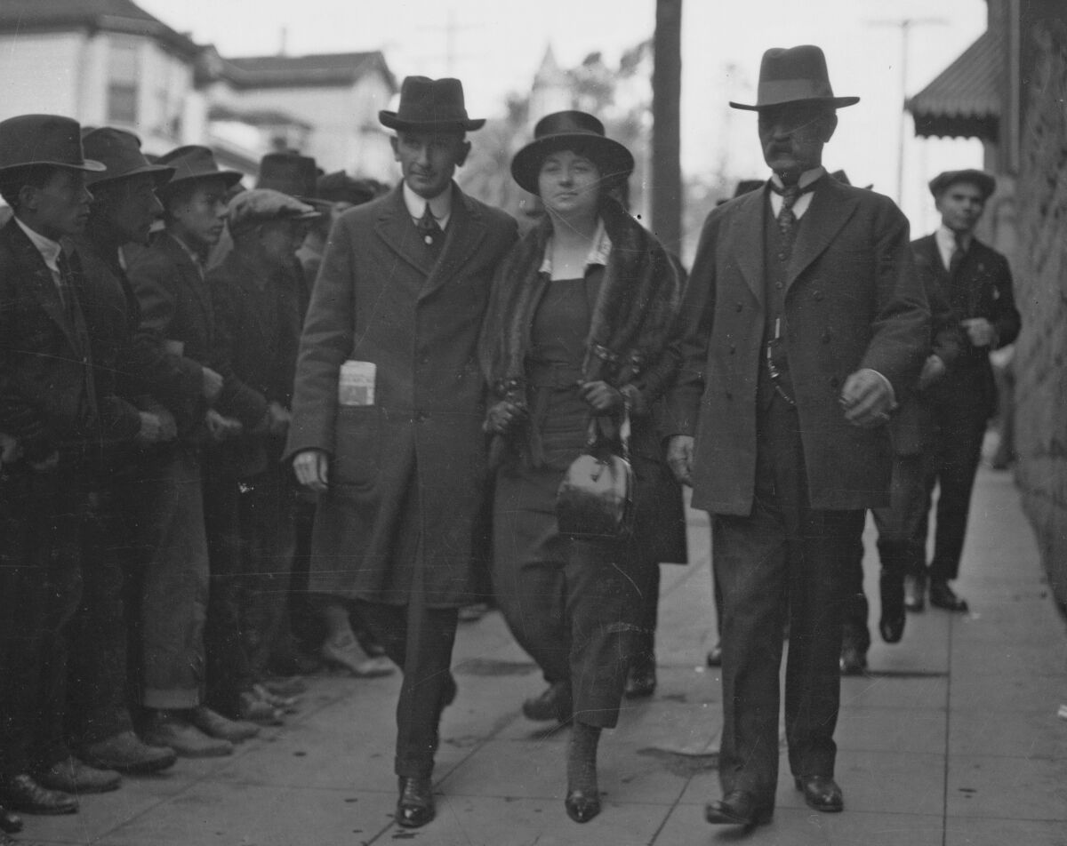 A woman walks between two men on a sidewalk lined by onlookers. Everyone has on hats; most men are in ties.
