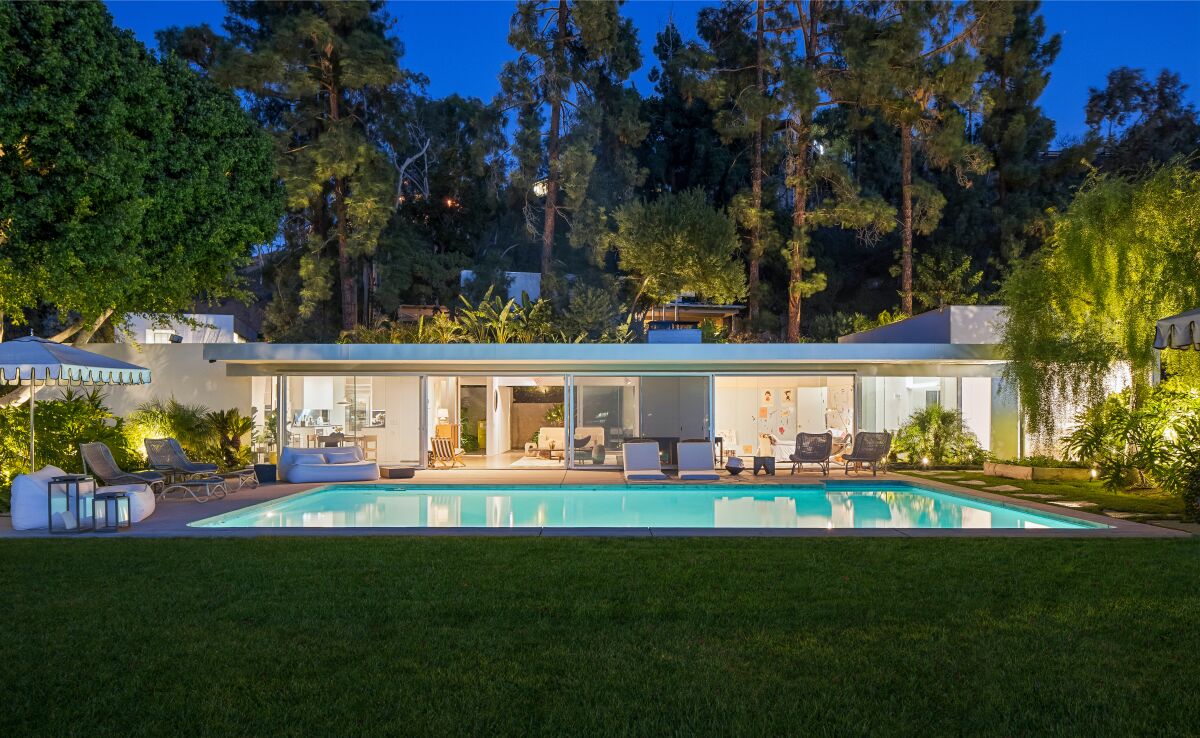 A long, low home with glass windows in back of a rectangular pool at dusk.
