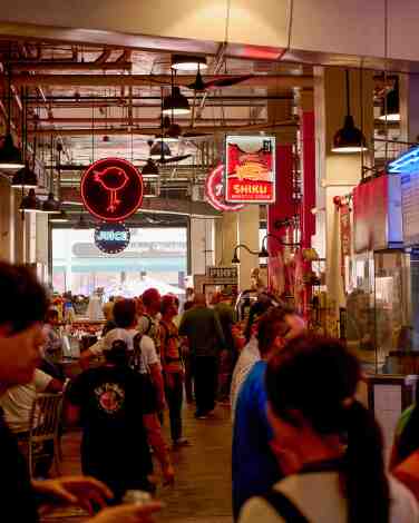 People walk in an open food market building under hanging signs