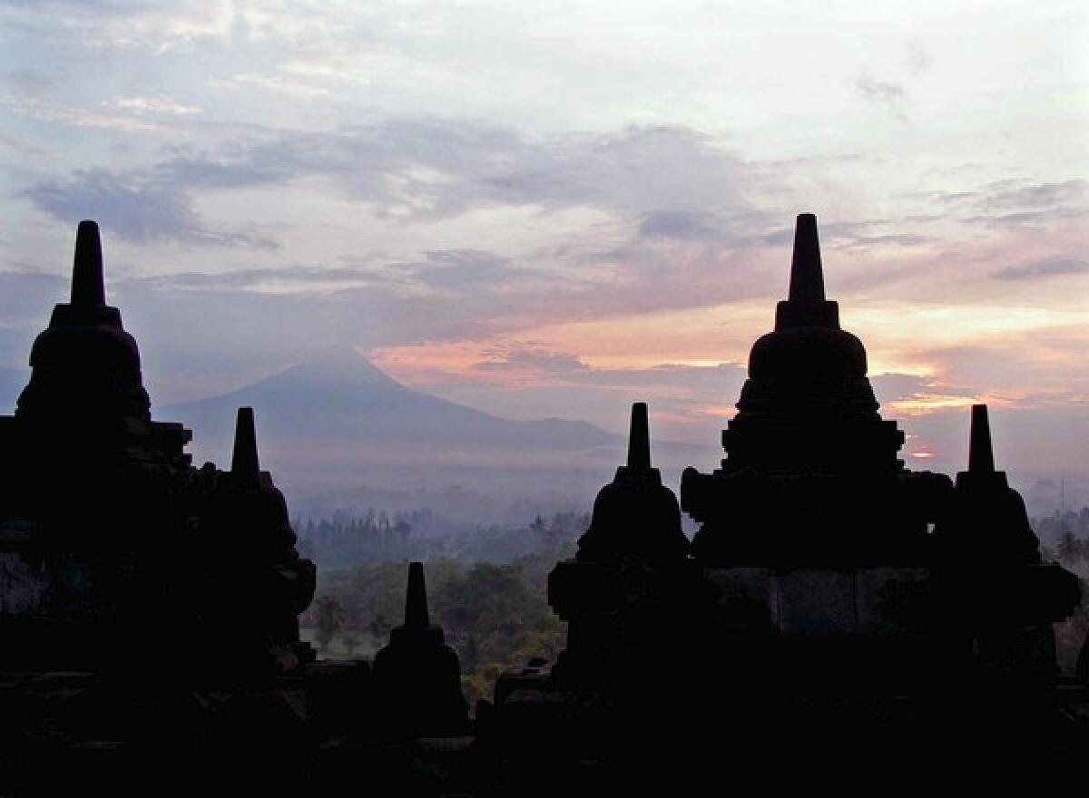 Mt. Merapi rises in the distance behind the forest of stupas at Borobudur, Indonesia. The stupas are perforated to allow glimpses at the Buddha figures inside.