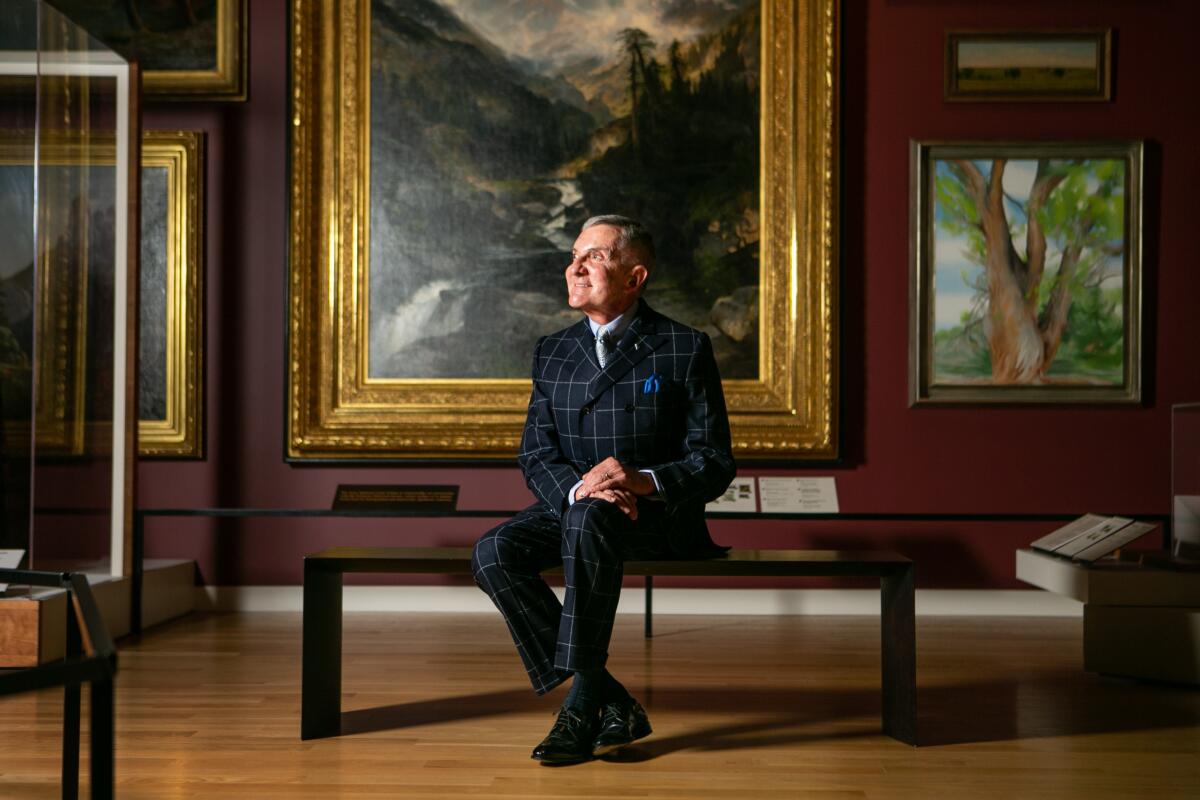 Rick West, in a checked suit, sits on a bench before a wall covered in landscape paintings in a museum gallery