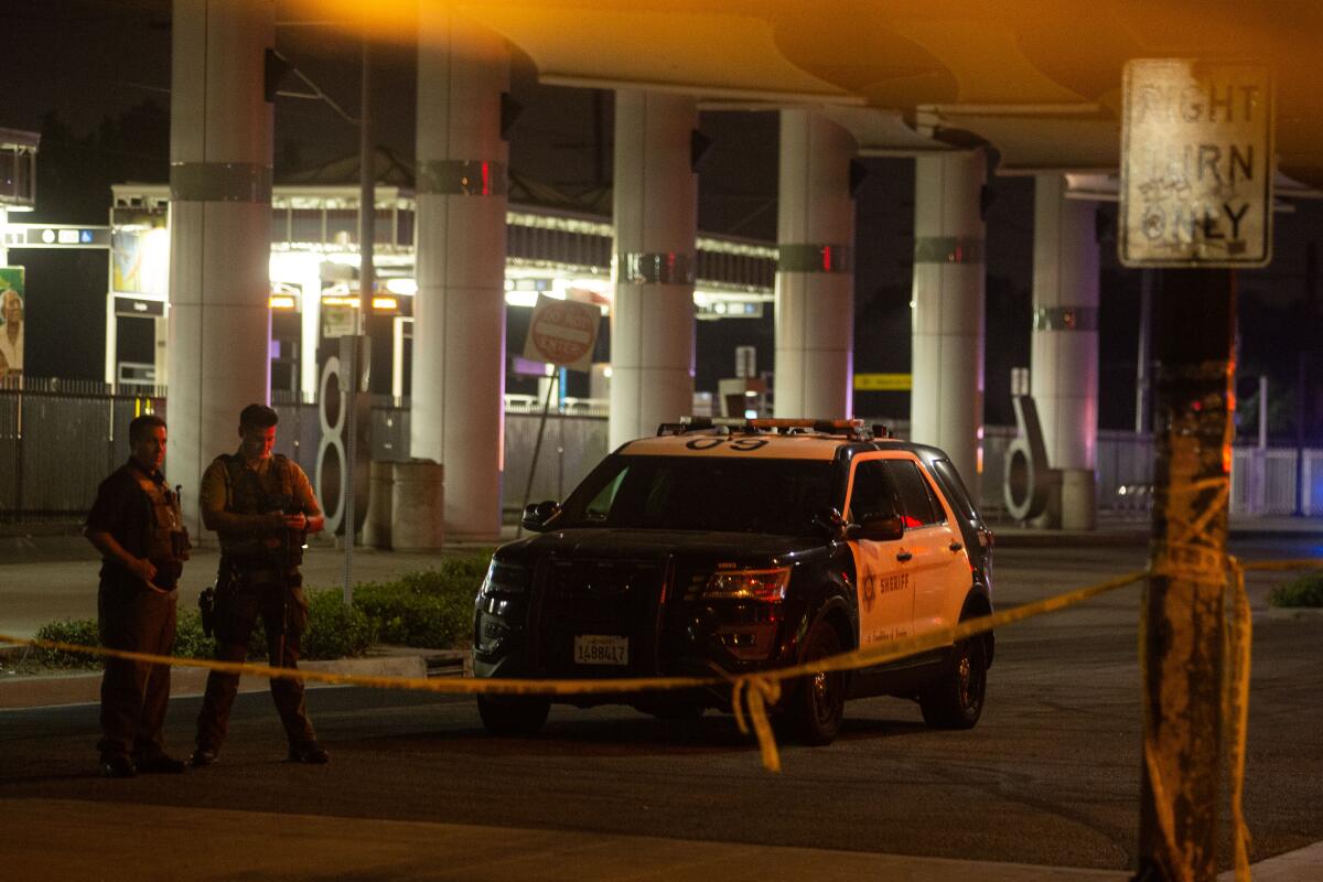 Two law enforcement officers in uniform stand next to a sheriff's SUV behind crime scene tape