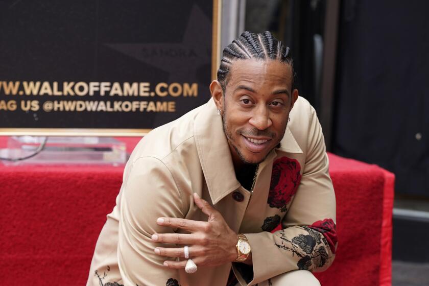 Ludacris, wearing a tan coat and pants, poses with hand resting on his star on the Hollywood Walk of Fame. 