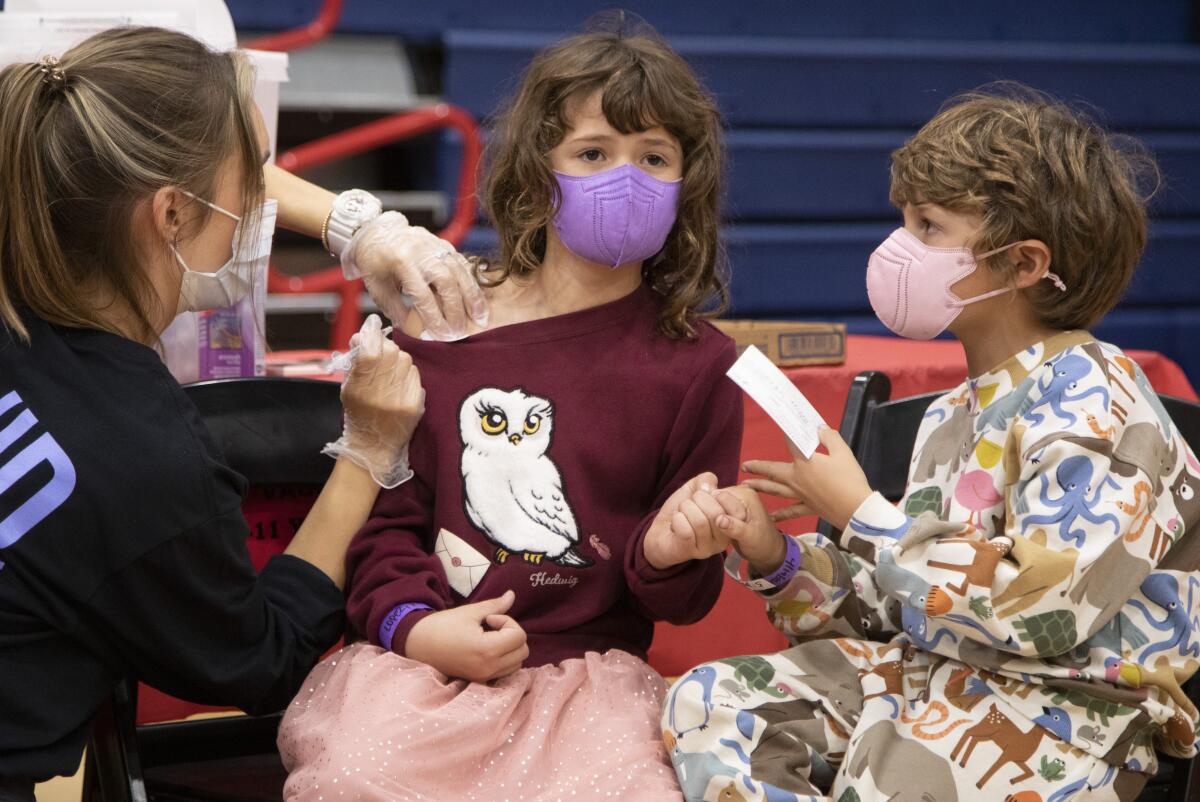 Two masked children hold hands as one of them gets a COVID vaccine.