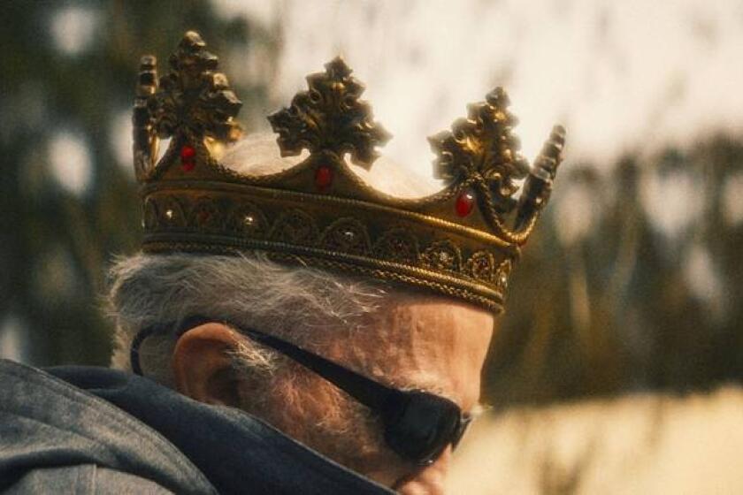 A gray haired man wears a crown and wrap-around dark sunglasses.