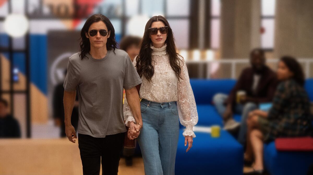 A man and woman in sunglasses walk through an office space holding hands in a scene from "WeCrashed."