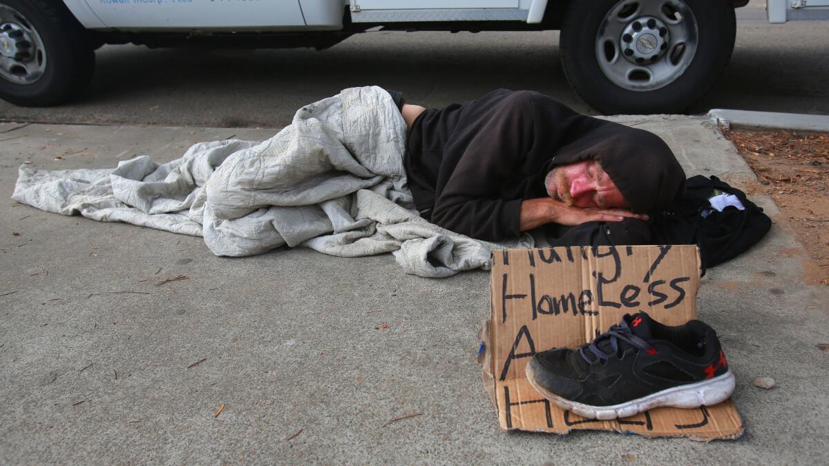 A man named Jerry sleeps on the sidewalk with nothing more than a blanket and a worn cardboard sign, as the morning dawns on downtown San Diego.