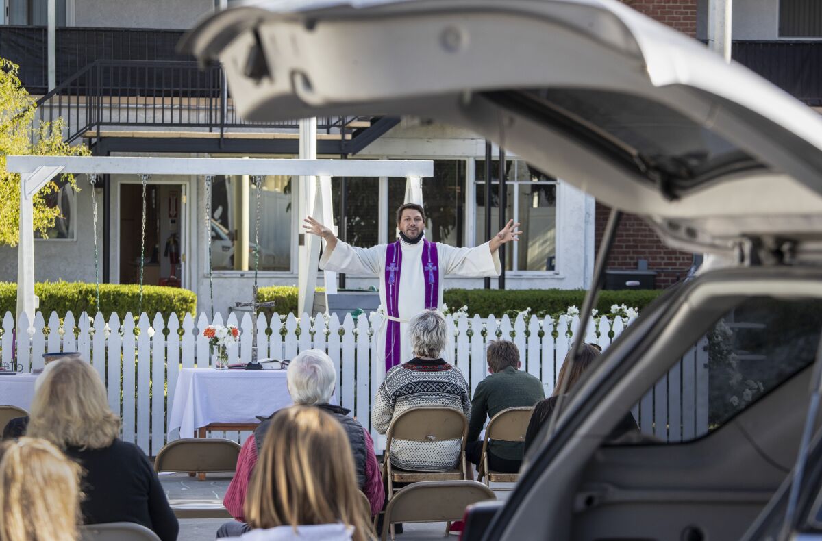 Framed by an open car trunk, people watch a minister lead a service in a parking lot.