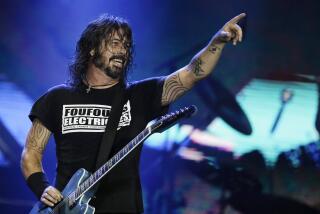 Dave Grohl in a black t-shirt on a stage pointing his left arm in the air and holding a guitar