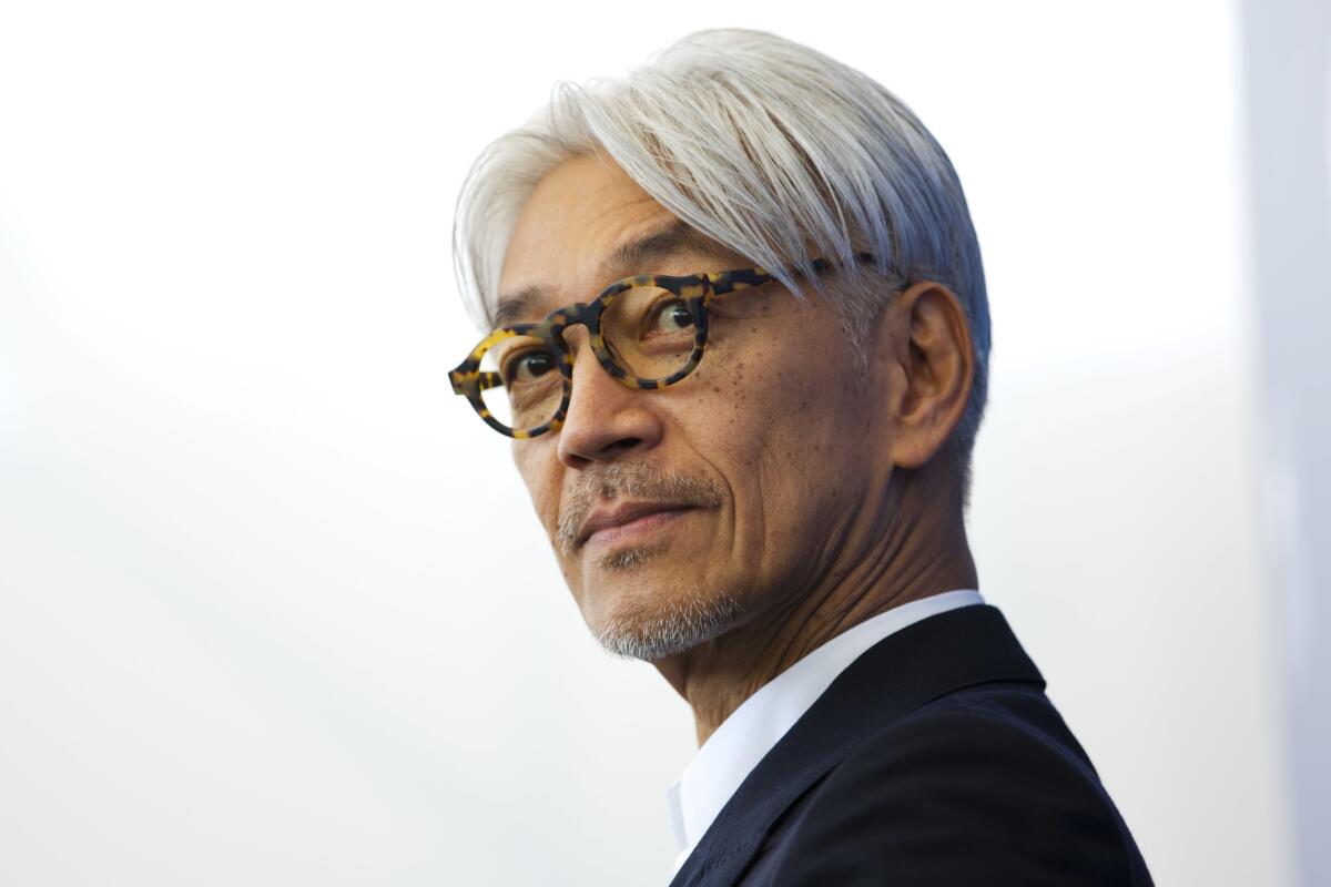 Ryuichi Sakamoto, in white shirt and black suit jacket, wearing tortoise shell glasses, looks over his left shoulder.