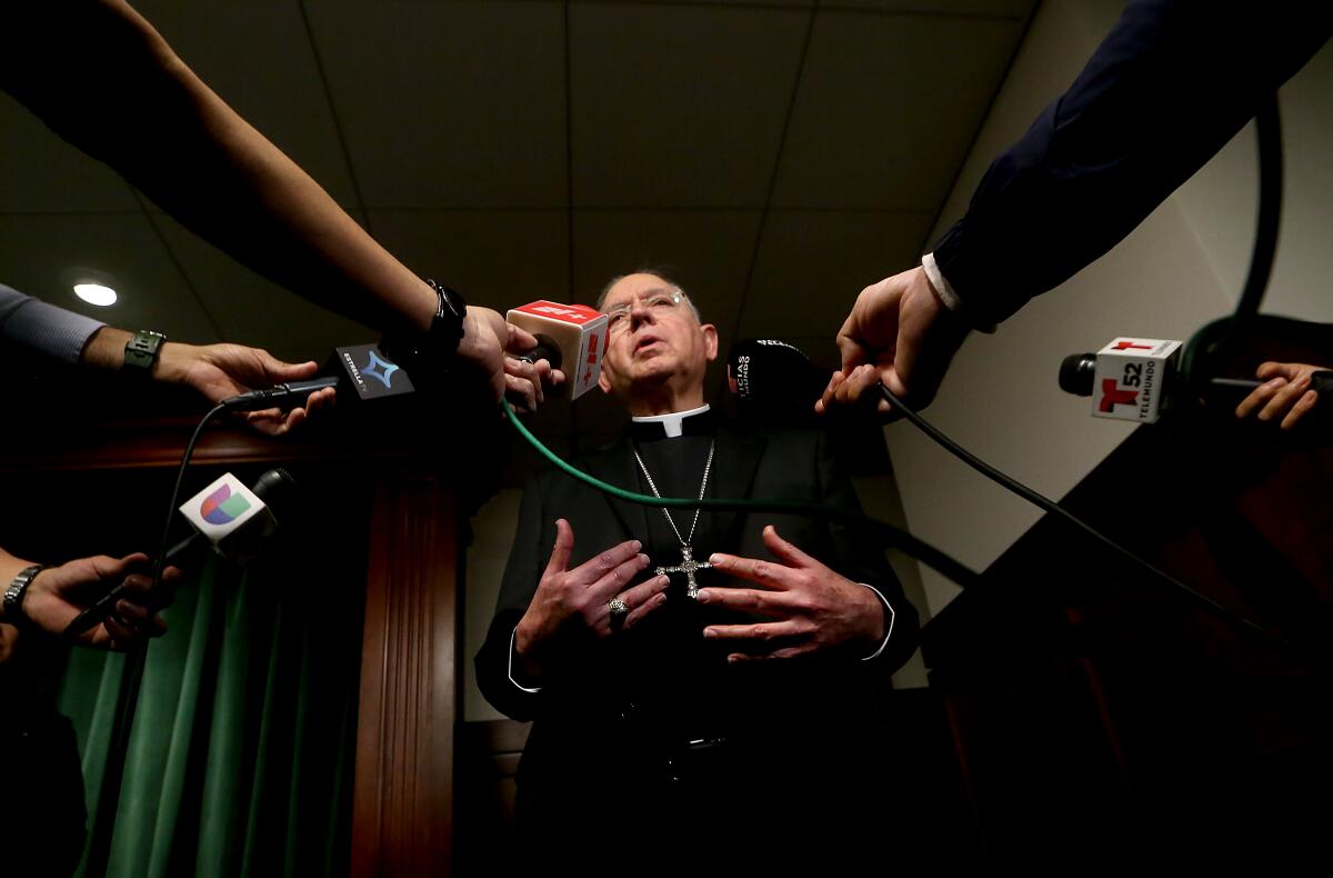 Seen from a low-angle view, a man wearing a black priest robe and a silver Christian-cross necklace speaks to reporters. 