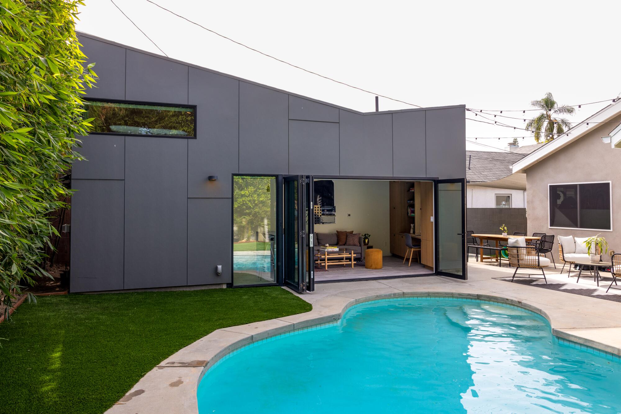 An ADU with a trapezoid-shaped roof sits behind a pool and outdoor furniture. 