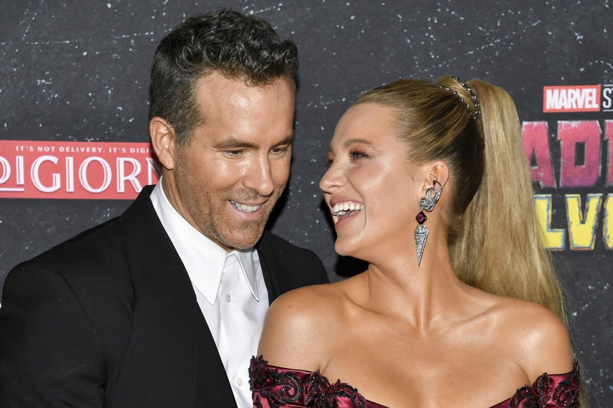 Ryan Reynolds wears a suit while his wife Blake Lively holds his hand and laughs in a red jumpsuit