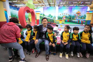 A man in a dark jacket sits, flanked by schoolchildren, in a colorful indoor setting