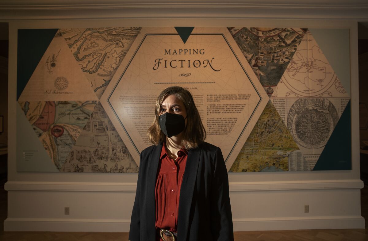 A woman donning a black mask and red blouse stands in front of an exhibition entrance. 