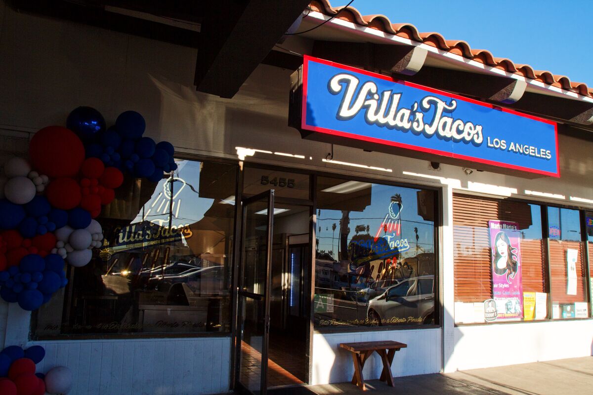 An exterior photo of a restaurant space in the strip mall. A blue-and-read sign reads "Villa's Tacos Los Angeles" in cursive