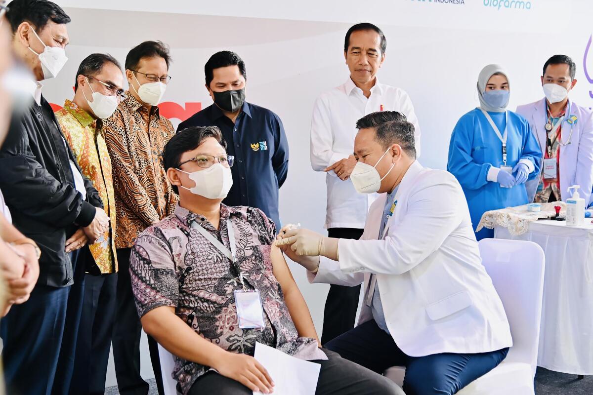 A group of people stand and watch as a seated man receives a shot in his left arm 