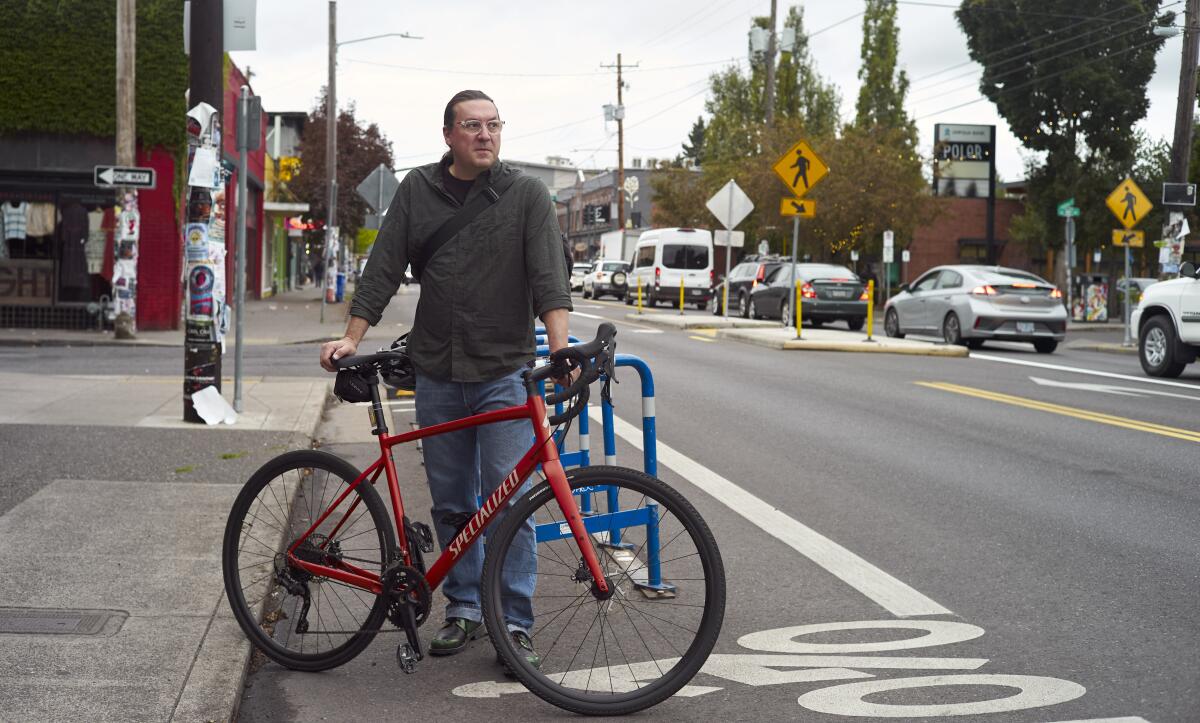 A photograph of a man standing with a bike on the side of a street. 