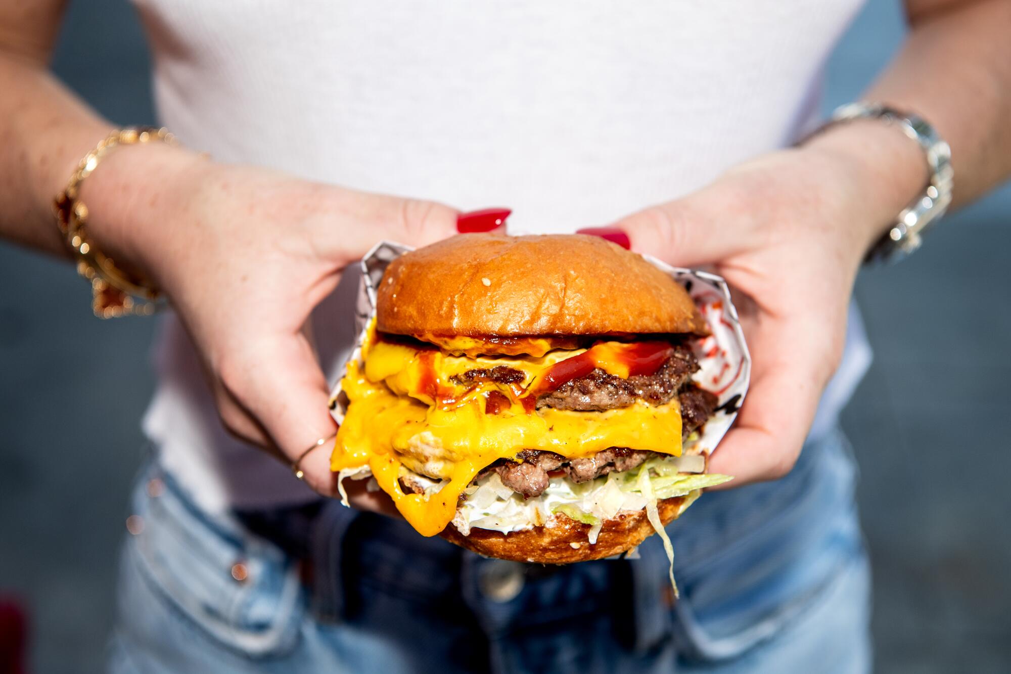 A closeup photo of a woman's hands holding a double cheeseburger oozing cheese and ketchup.