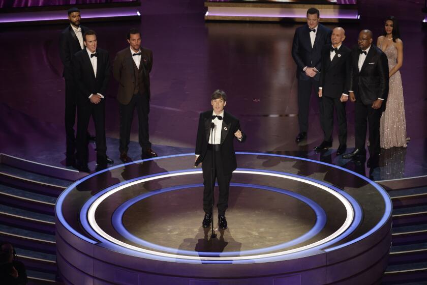 Cillian Murphy wears a suit while accepting an Oscar on a stage surrounded by other men in suits.