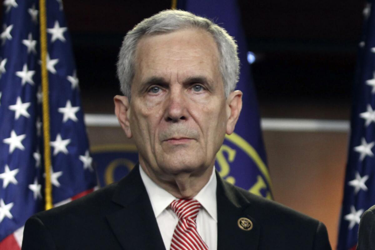 Lloyd Doggett from the shoulders up in a suit jacket and red-and-white-striped tie, in front of U.S. House and American flags