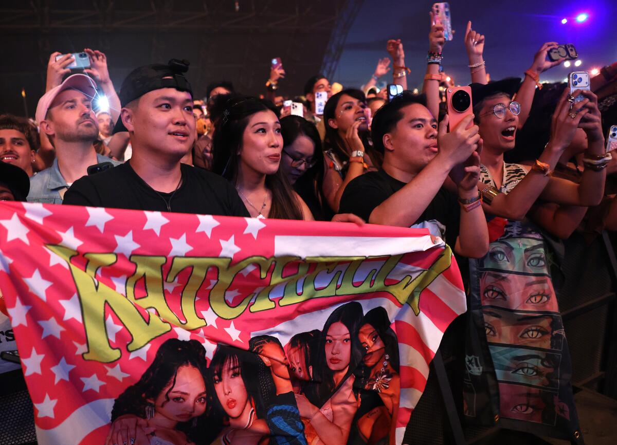 Fans watch Katseye perform and one holds a flag with the group's name and image