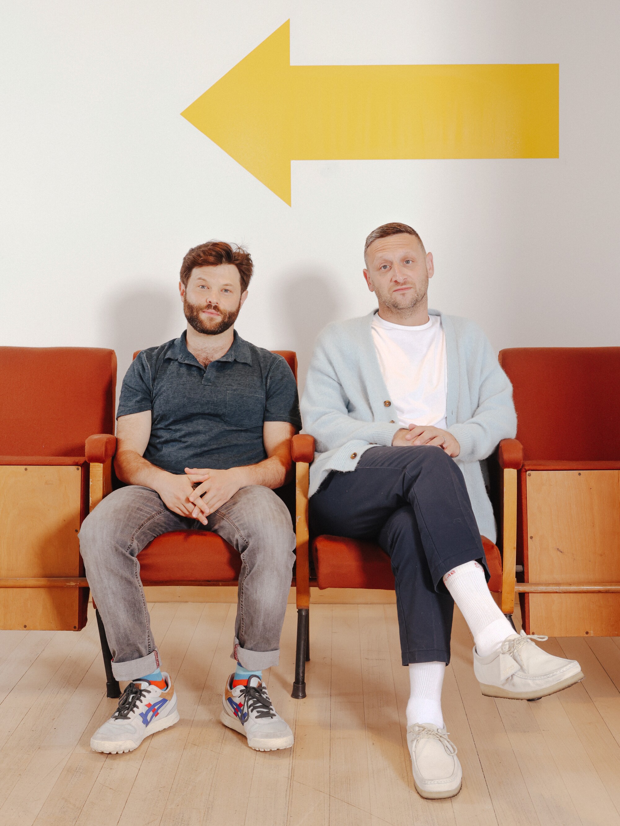 Two men are sitting on chairs, behind them a large yellow arrow on the wall.