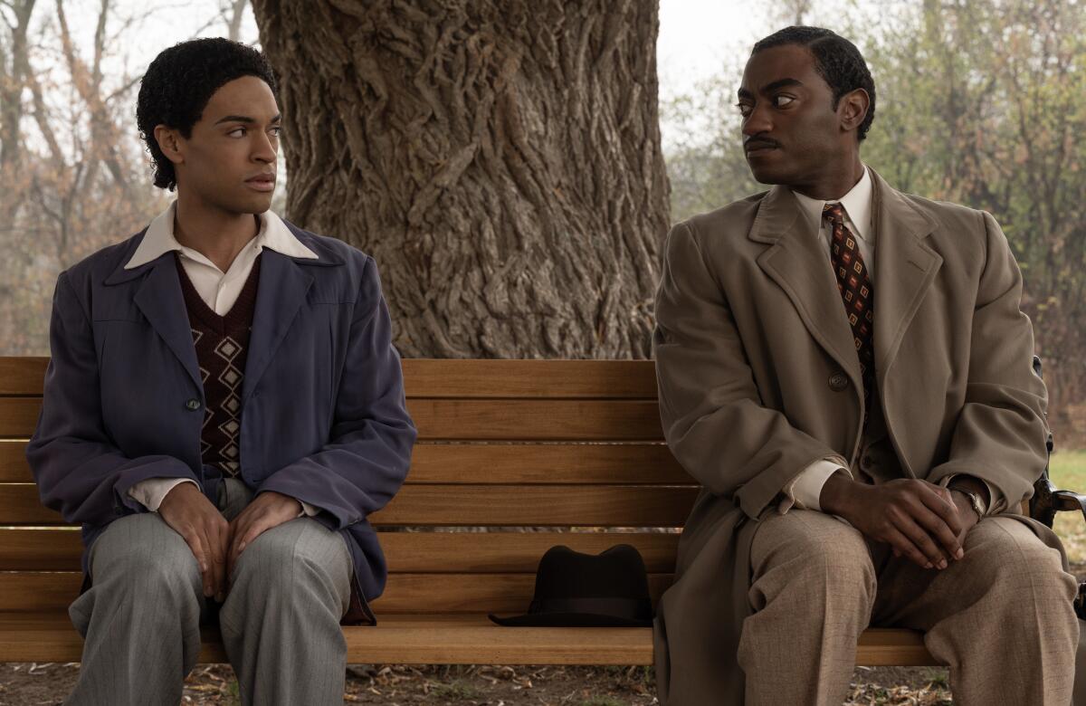 Two men sit on a bench outdoors, looking at each other.