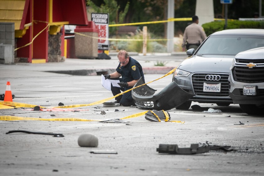 An officer kneels on the ground next to vehicles and debris lying in the street.
