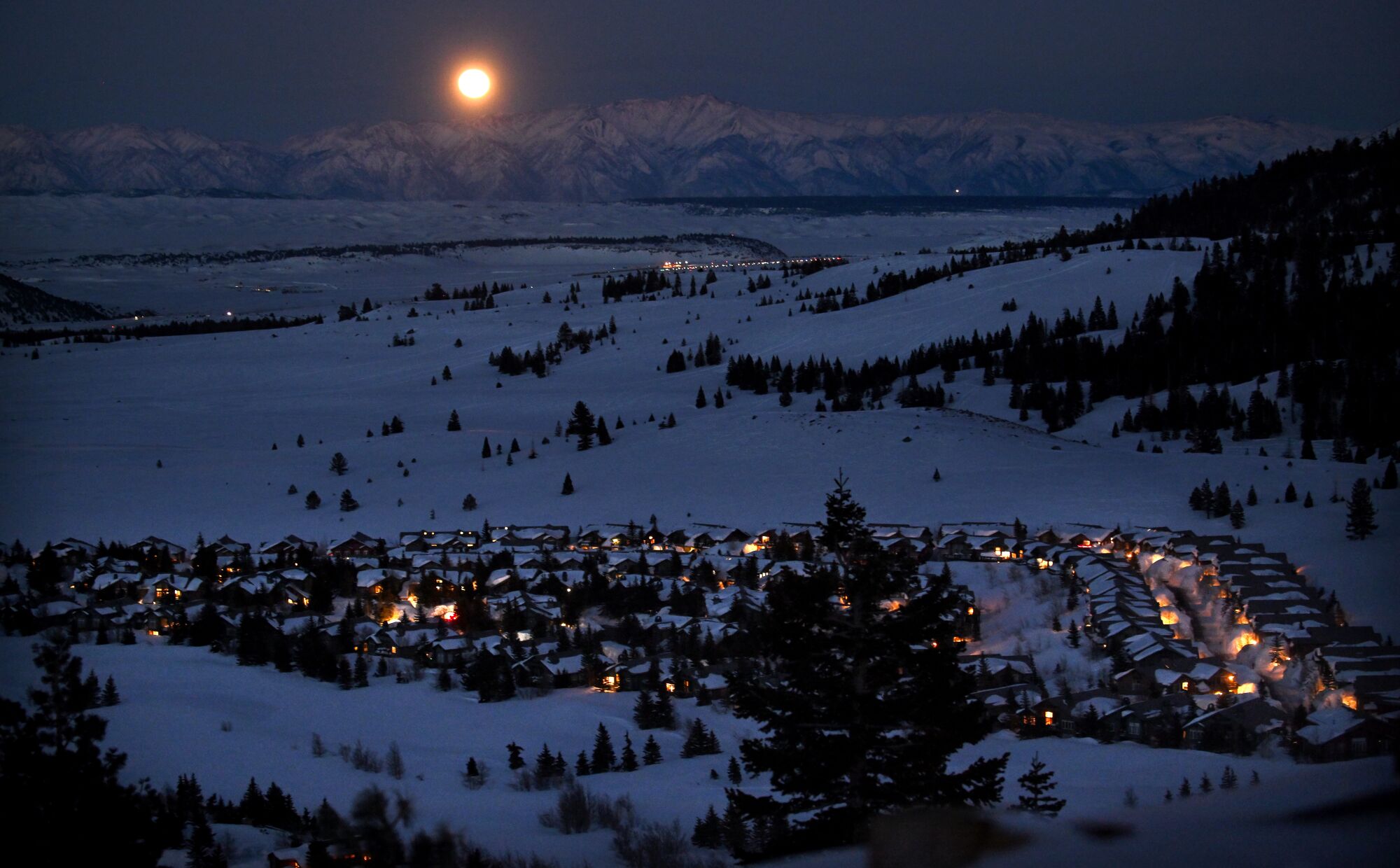 A full moon rises over a valley with a village at night, with mountains in the background