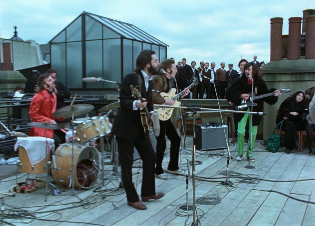 The Beatles play their instruments on top of a roof with a small crowd watching.