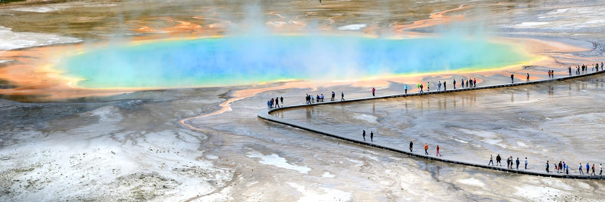 Hikers file past a spring of rainbow hues. Grand Prismatic Spring, Yellowstone National Park, Wyo. 