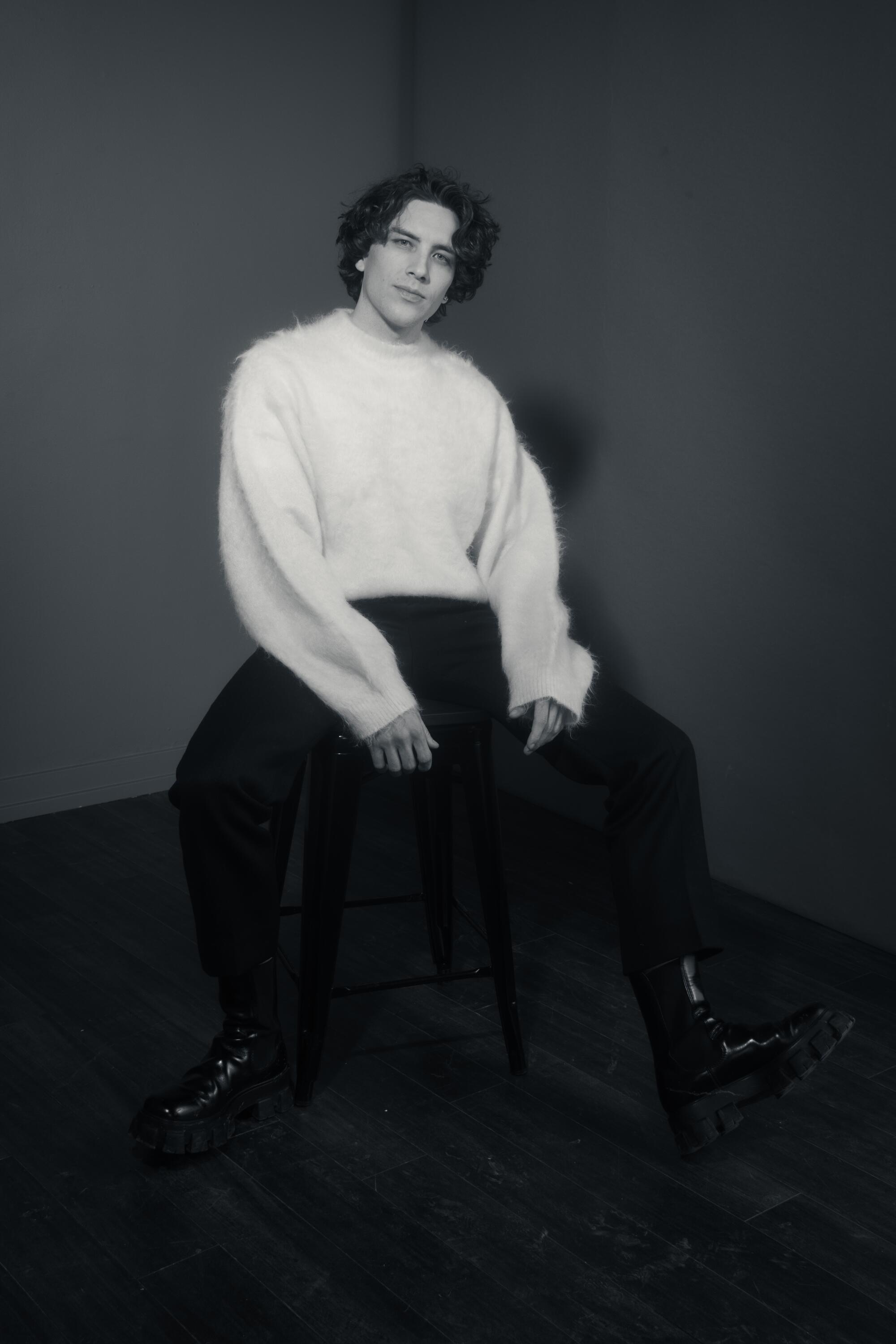 Cody Fern in a fuzzy sweater and dark pants sits on a stool.