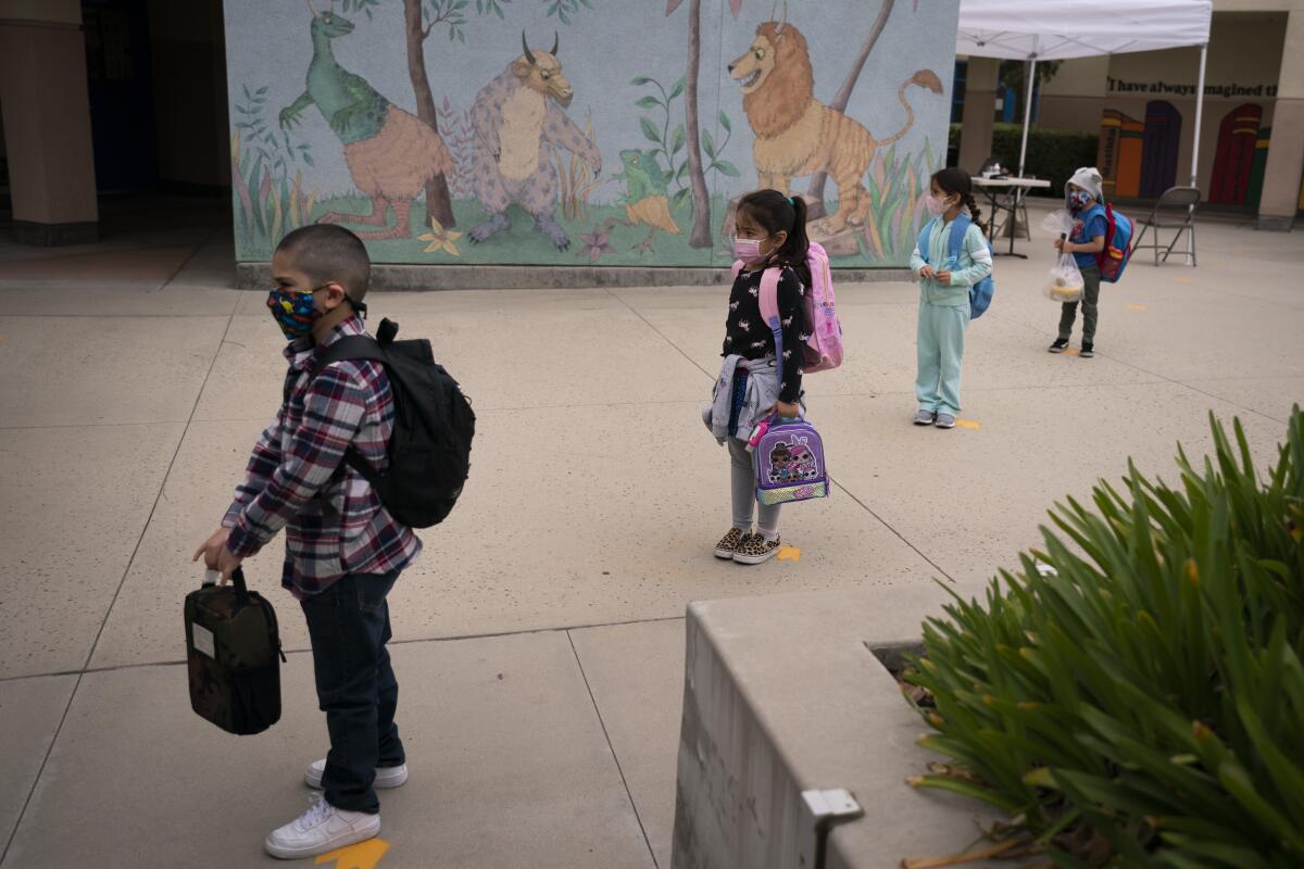 Kids with backpacks and lunch boxes stand socially distanced at a school