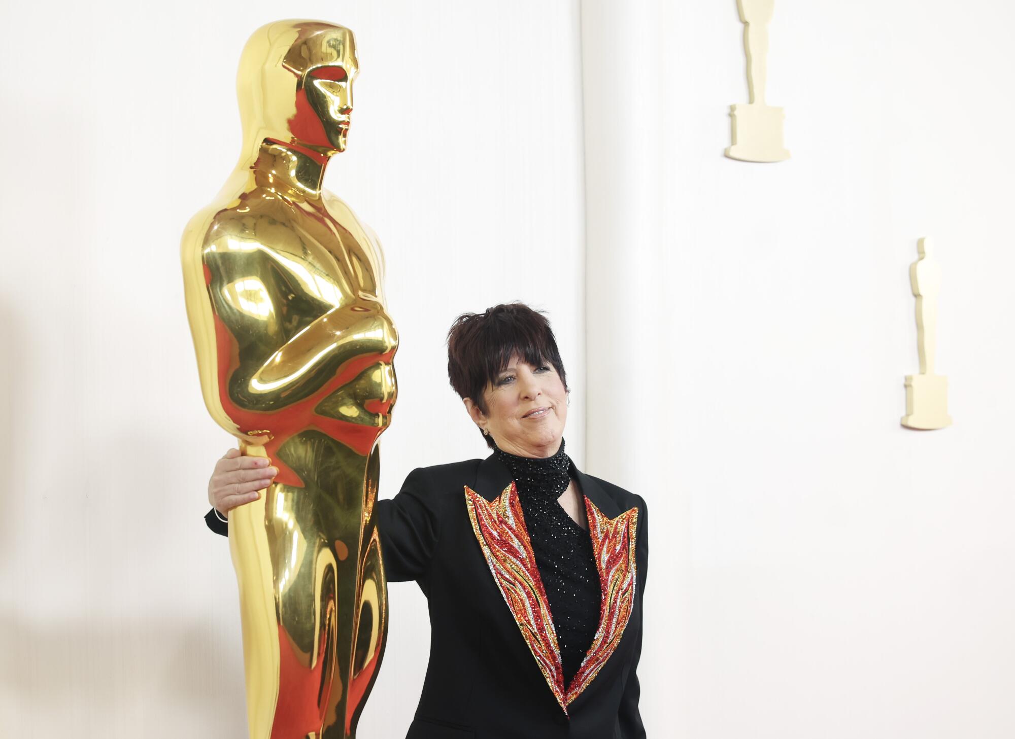 Diane Warren, in a black suit with a fire design on the lapels, stands next to an Oscar statue. 